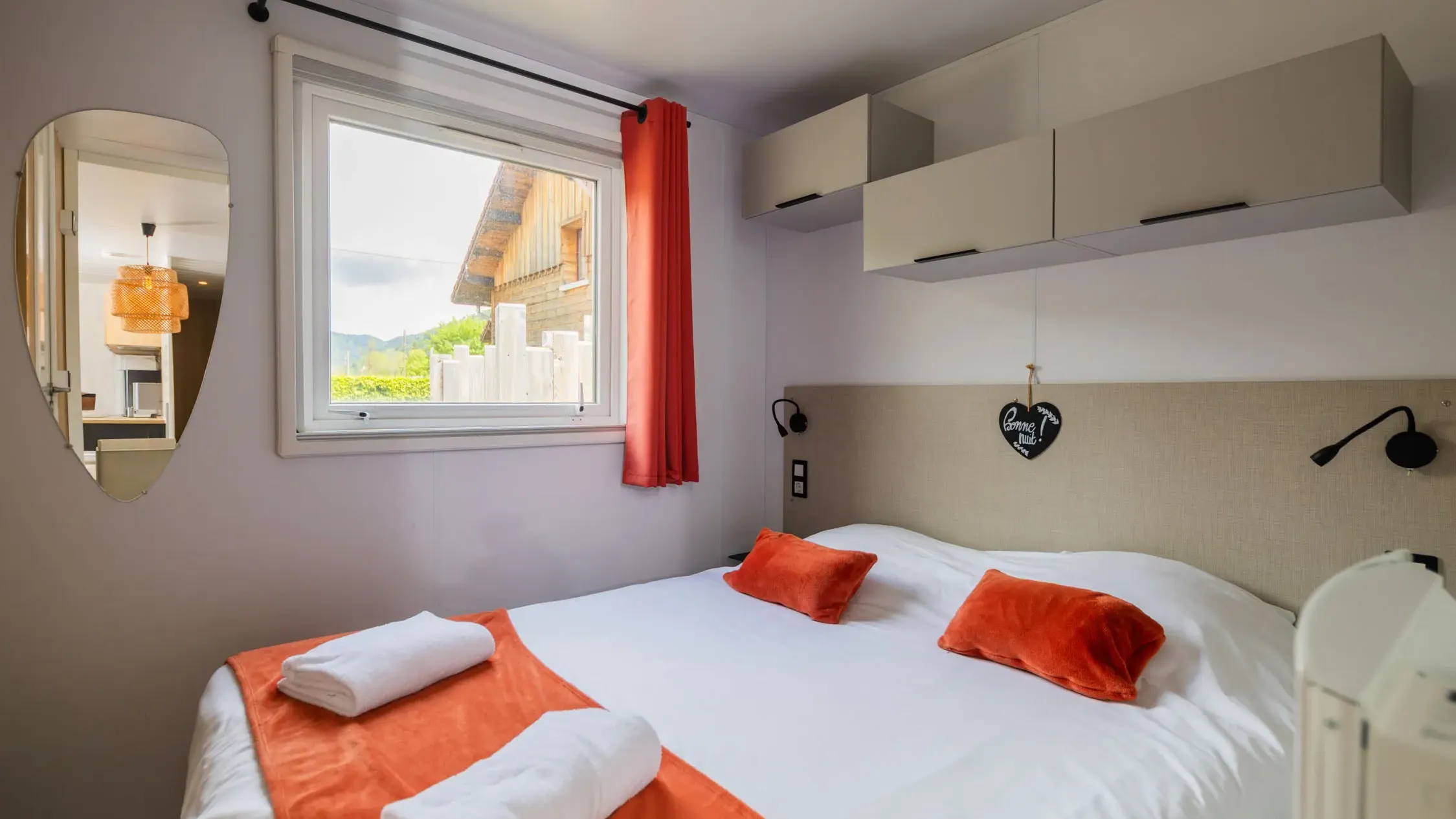Room with a double bed, white and orange towels, a window with a red curtain, wall shelves, and a heart-shaped mirror