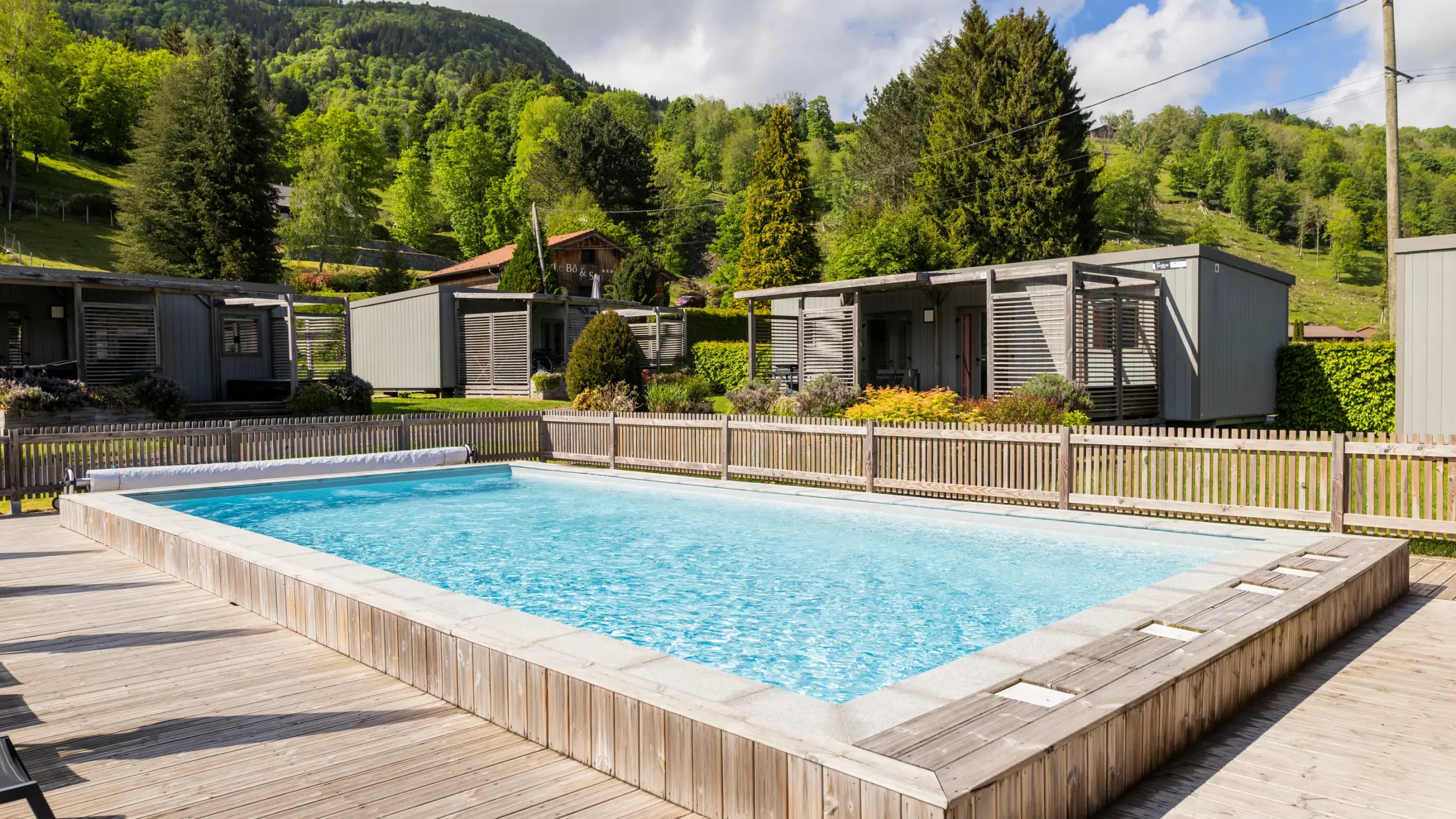Rectangular swimming pool surrounded by a wooden deck with wooden cabins behind and a green hill in the background.