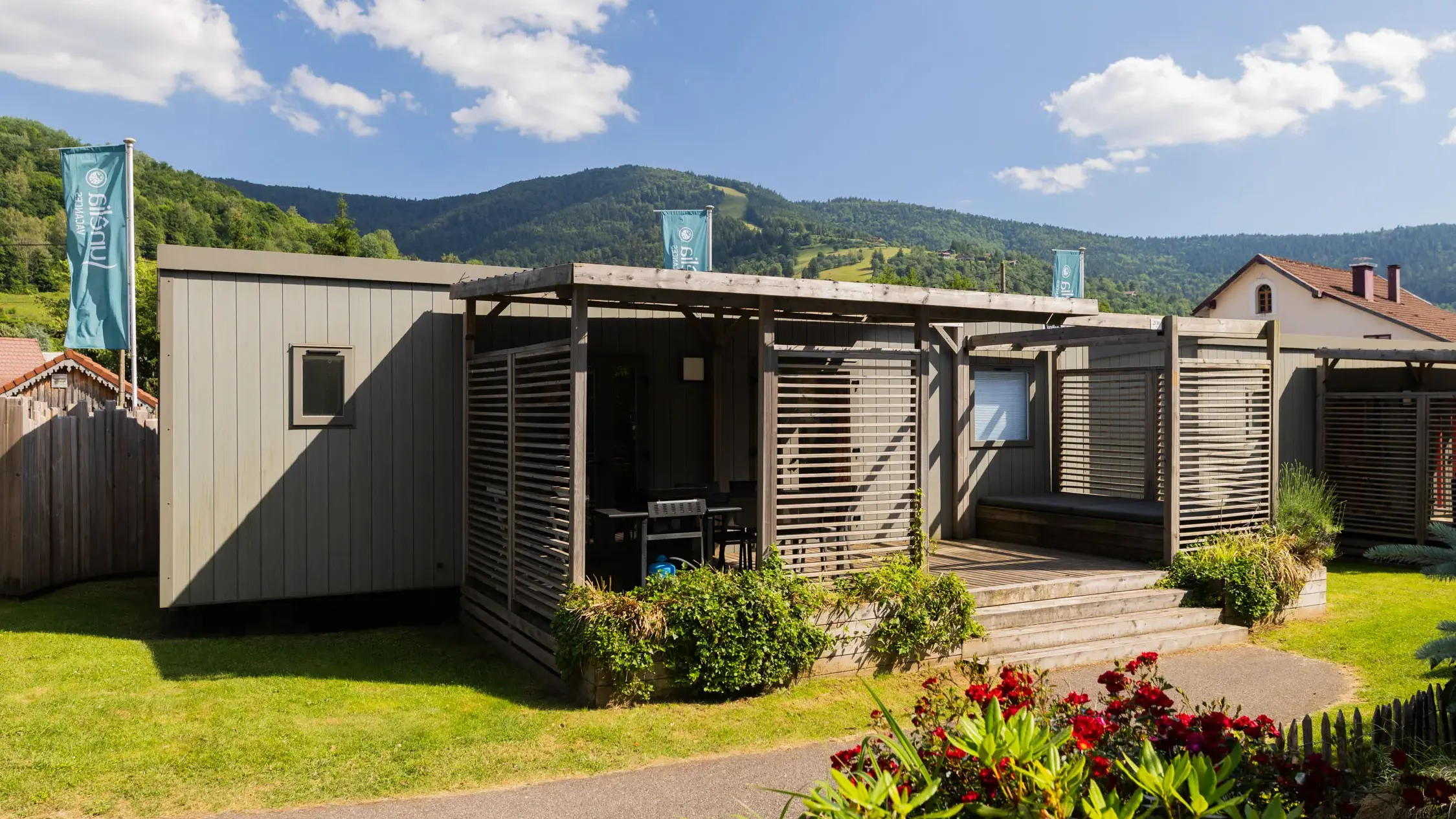 Small wooden cabin with terrace and plants, located in a garden with mountains in the background